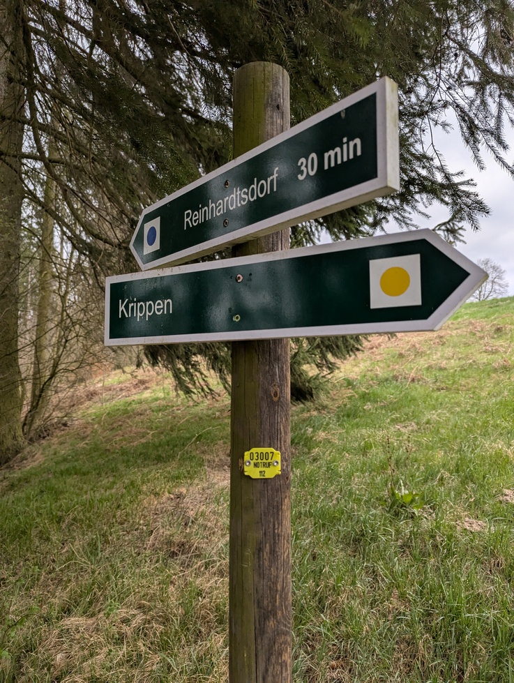 Wegweiser Reinhardtsdorf Krippen mit gelben Notrufpunktschild vor grünem Hügel