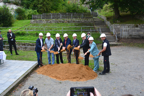 mehrere Personen mit Bauhelm und Spaten vor Erdhügel